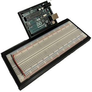 May include: An Arduino Uno microcontroller board with a breadboard, enclosed in a black case. The breadboard has red and blue lines, and a few wires are connected. The Arduino board is green with various electronic components.