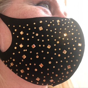 Puede incluir: Mascarilla facial negra adornada con pedrería dorada de varias formas y tamaños. La mascarilla está hecha de un material suave y ajustado, diseñada para cubrir la nariz y la boca.