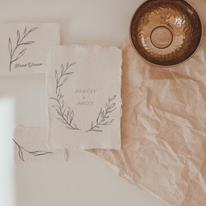 May include: A collection of handmade paper cards with botanical illustrations and handwritten text. The cards feature delicate line drawings of leaves and floral wreaths in gray ink. A hammered brass bowl sits on a textured, tan surface.
