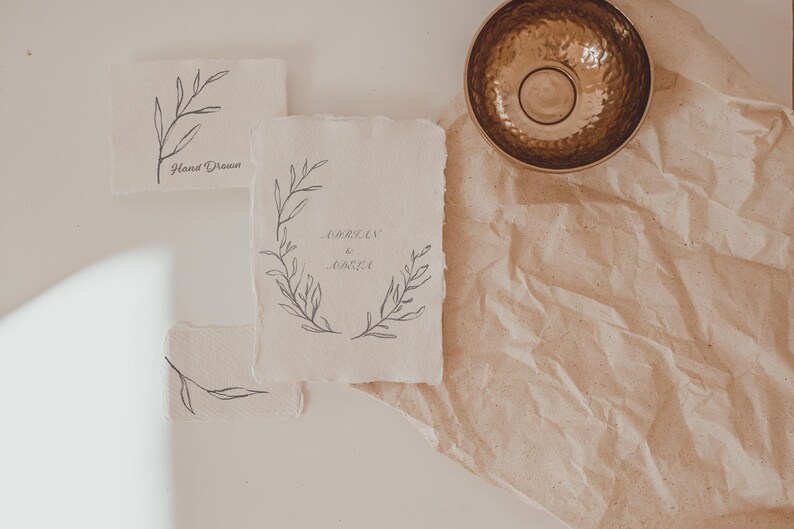 May include: A collection of handmade paper cards with botanical illustrations and handwritten text. The cards feature delicate line drawings of leaves and floral wreaths in gray ink. A hammered brass bowl sits on a textured, tan surface.