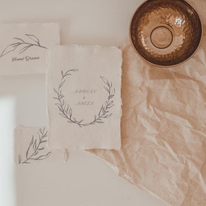 May include: A collection of handmade paper cards with botanical illustrations and handwritten text. One card features a wreath design with the names "Adrian & Odela". A hammered metal bowl sits on a textured paper background.