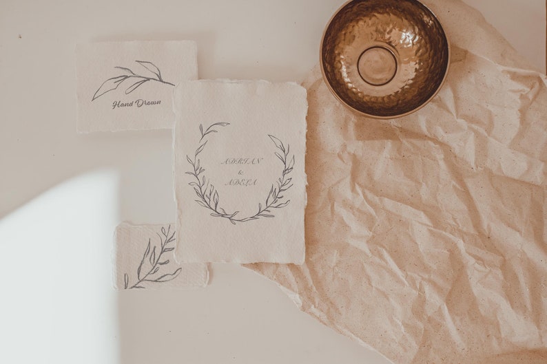 May include: A collection of handmade paper cards with botanical illustrations and handwritten text. One card features a wreath design with the names "Adrian & Odela". A hammered metal bowl sits on a textured paper background.
