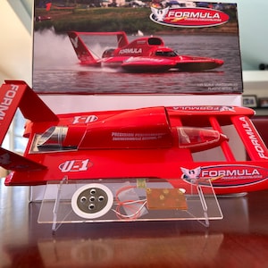 May include: A red Formula model boat with the text "FORMULA" on the side and rear fins. The model boat is on a clear plastic stand. The box behind the boat shows a picture of the boat in action.