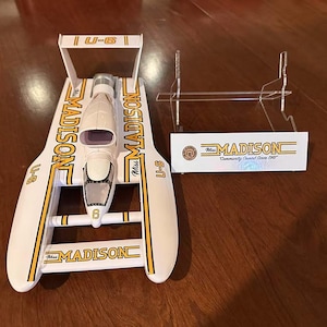 May include: A white and yellow model hydroplane boat with the name "Madison" printed on the sides. The boat has the number "6" on the front and "U-6" on the sides. A clear display stand and a sign with the "Miss Madison" logo are also present.