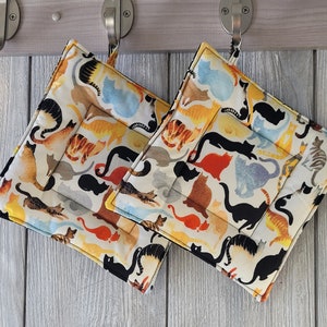 May include: Two white cotton pot holders with a colorful cat print. The pot holders are hanging on hooks.