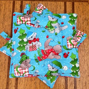 May include: Two blue and white Christmas-themed pot holders with a cat and mouse design. The pot holders have a red and white checkered border and a red hanging loop.