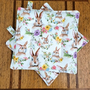 May include: Two white pot holders with a floral pattern featuring brown bunnies and colorful flowers. The pot holders are made of fabric and have a quilted texture.