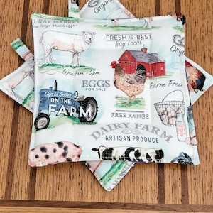May include: A white and blue farm-themed pot holder with images of a sheep, chicken, pig, and cow. Text includes "Eggs for Sale", "Dairy Farm", and "Fresh is Best Buy Local". The pot holder has a square shape with a loop for hanging.