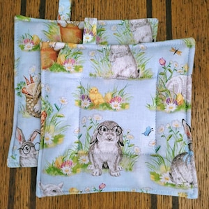 May include: Two square, light blue pot holders with an Easter theme. The fabric features illustrations of bunnies wearing glasses, chicks, eggs, flowers, and butterflies. Each pot holder has a loop for hanging.