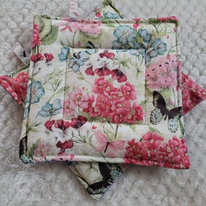 May include: Two floral print pot holders with butterflies and pink flowers. The pot holders are quilted and have a light beige background.