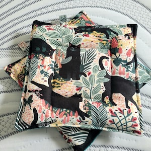 May include: Two black cat-themed pot holders with a green, white, and pink floral pattern. The pot holders are made of quilted fabric and have a loop for hanging.