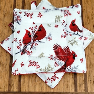 May include: Two white square pot holders with a red cardinal bird and berry design. The pot holders have a red trim and a loop for hanging. The design features red cardinals perched on branches with red berries.
