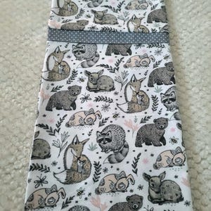 May include: A white fabric item featuring a repeating pattern of woodland animals, including foxes, raccoons, bears, and deer. The design incorporates floral and leaf elements in pink and green. A gray polka-dot ribbon accents the top.