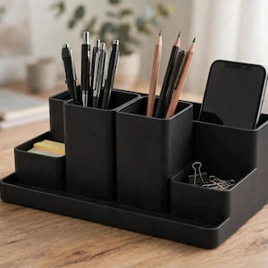 Peut inclure: Un organisateur de bureau noir mat avec plusieurs compartiments de hauteurs variées. Il contient plusieurs stylos, crayons, un smartphone, une pile de notes adhésives jaunes et des pinces à dessin en métal argenté sur une base rectangulaire.