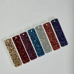 May include: Seven rectangular glitter tags in a range of colours: gold, red, purple, teal, silver, orange, and navy. Each tag has a small hole at the top for hanging. The tags are arranged diagonally against a white background.