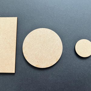 May include: Three unfinished wooden shapes: a rectangle, a large circle, and a small circle. The shapes are light brown and have a smooth, unpainted surface. They are arranged on a dark grey background.