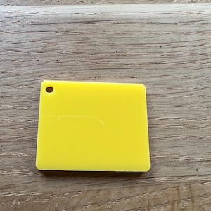 May include: A vibrant yellow, rectangular acrylic tag with a rounded corner and a small hole at the top. The tag is smooth and likely a sample or blank for personalisation. It rests on a light brown wooden surface.