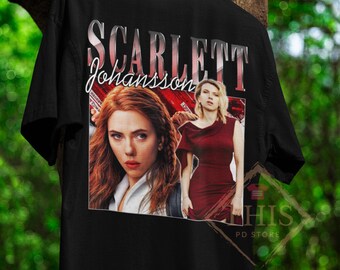 May include: Black t-shirt featuring a graphic of Scarlett Johansson. The design includes her name in a stylized font and two images of the actress. The shirt is hanging on a wooden hanger against a blurred green background.