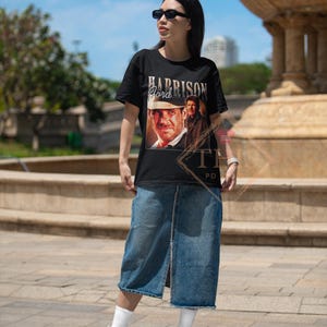 May include: A black t-shirt with a graphic print of Harrison Ford as Indiana Jones. The shirt has a crew neck and short sleeves. The model is wearing a denim skirt and white sneakers.