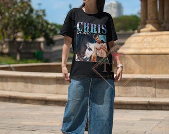 May include: Black t-shirt featuring the name "Chris Evans" in a stylized font, with images of the actor. The person is wearing blue denim culottes and white sneakers. The background includes a fountain and a blue sky.
