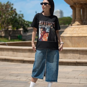 May include: A woman wearing a black t-shirt with a graphic of Benedict Cumberbatch as Doctor Strange. The shirt has the text "Benedict Cumberbatch" printed on it. She is also wearing a blue denim skirt and white sneakers.