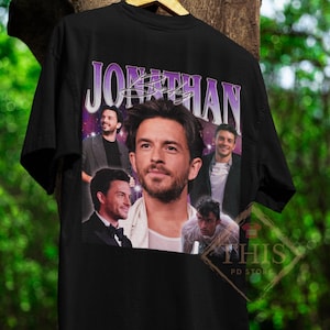 May include: Black t-shirt with a purple and white graphic print of a man's face and the word "JONATHAN" in a retro font.