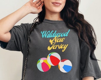 May include: A gray cropped t-shirt with the text "Wildwood New Jersey" and a graphic of beach balls. The shirt is being worn by a person with long brown hair and a white pleated skirt.
