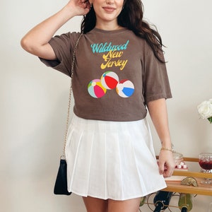 May include: A brown t-shirt with a graphic of beach balls and the text "Wildwood New Jersey" in a retro font. The person is wearing a white pleated skirt.