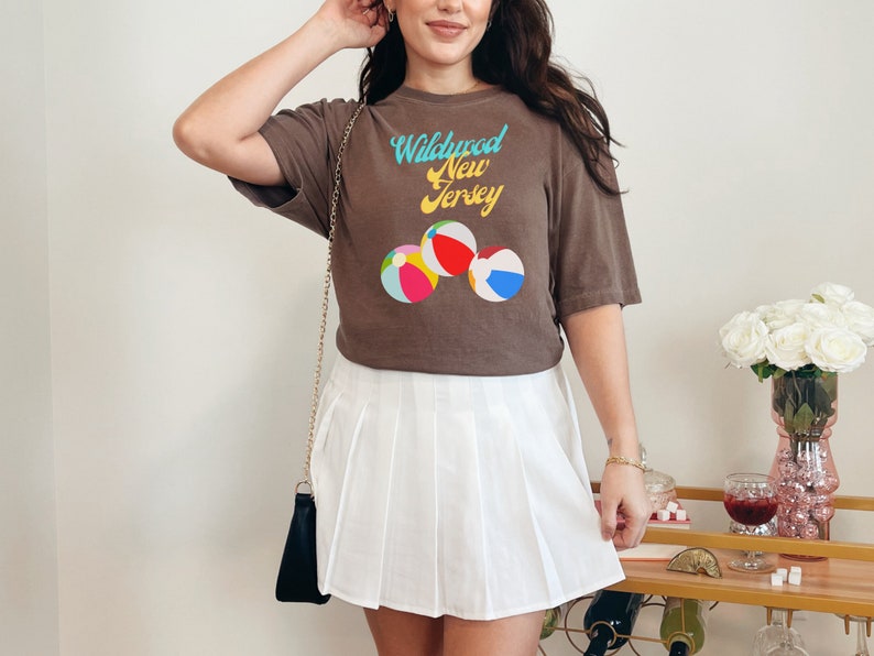May include: A brown t-shirt with a graphic of beach balls and the text "Wildwood New Jersey" in a retro font. The person is wearing a white pleated skirt.