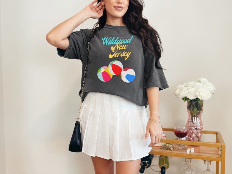 May include: A gray cropped t-shirt with the text "Wildwood New Jersey" and a graphic of beach balls. The shirt is being worn by a person with long brown hair and a white pleated skirt.