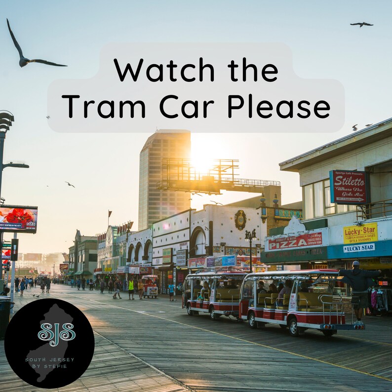 May include: A view of the Atlantic City boardwalk with a tram car driving down the middle of the boardwalk. The tram car is red and white with yellow accents. The tram car is full of people. The boardwalk is made of wood and there are buildings on either side of the boardwalk. The sky is blue and there are clouds in the sky. The sun is shining brightly. The text "Watch the Tram Car Please" is written in white letters on a gray background. The text "SJS South Jersey by Stefie" is written in white letters on a black background.