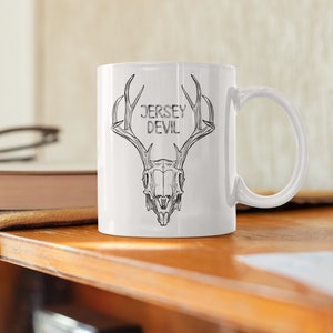 May include: White ceramic mug with a black and white illustration of a deer skull with antlers. The text "JERSEY DEVIL" is printed on the mug.