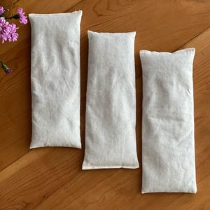 Op de afbeelding: Drie rechthoekige, lichtgrijze stoffen zakken liggen op een houten oppervlak. De zakken zijn gevuld en hebben een zacht, getextureerd uiterlijk. Een paar paarse bloemen zijn zichtbaar in de linkerbovenhoek.