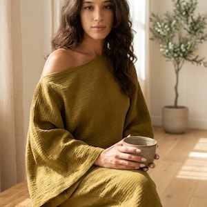 May include: An olive green long-sleeved top with a textured, crinkled fabric finish and a wide, off-the-shoulder neckline. The garment features loose, flowing sleeves and a relaxed fit, paired with matching olive green pants in the same fabric.