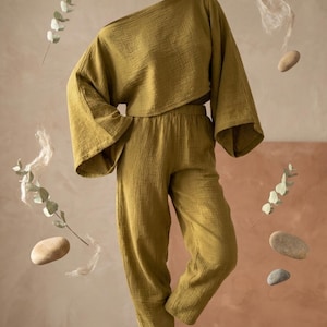 May include: An olive green two-piece set consisting of a loose-fitting, long-sleeved cropped top with wide sleeves and matching high-waisted trousers with a tapered leg. The fabric has a visible crinkled texture throughout the entire garment.