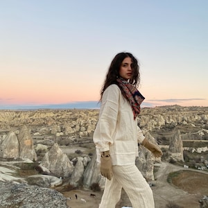 May include: A woman wearing a white linen pantsuit and brown leather gloves stands on a rocky cliff overlooking a landscape of rock formations. She is wearing a patterned scarf around her neck and has long brown hair.
