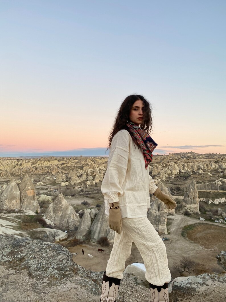 May include: A woman wearing a white linen pantsuit and brown leather gloves stands on a rocky cliff overlooking a landscape of rock formations. She is wearing a patterned scarf around her neck and has long brown hair.