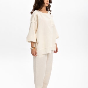 May include: A cream-coloured, loose-fitting, long-sleeved top and matching trousers. The top has a round neckline and a slightly gathered look. The trousers are wide-legged and have a relaxed fit.