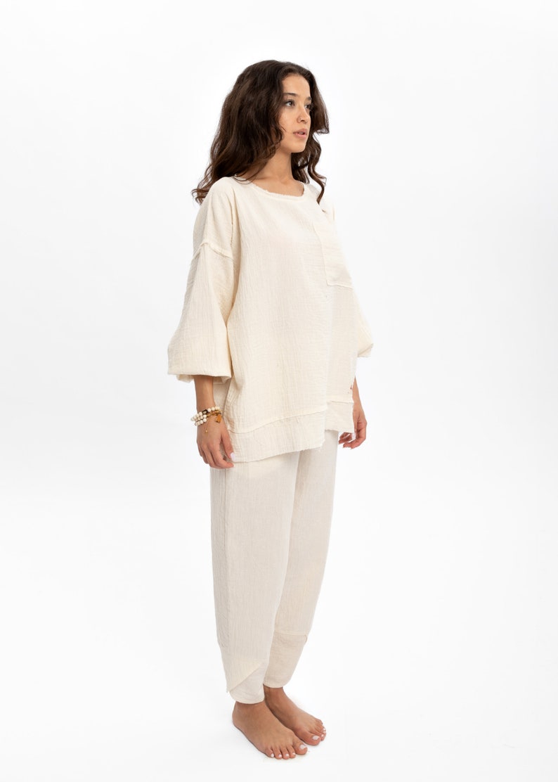 May include: A cream-coloured, loose-fitting, long-sleeved top and matching trousers. The top has a round neckline and a slightly gathered look. The trousers are wide-legged and have a relaxed fit.