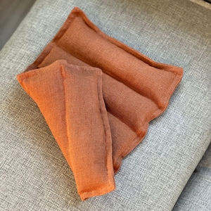 Könnte beinhalten: Ein gefaltetes, rostfarbenes Heizkissen aus Leinen. Das Kissen ist rechteckig mit einem gewellten Rand und ist zur Hälfte gefaltet. Der Stoff hat ein strukturiertes Aussehen. Das Kissen liegt auf einer grauen, gepolsterten Oberfläche.