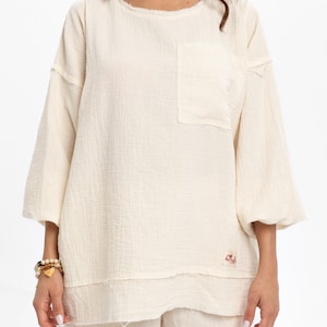 May include: A white, loose-fitting, 3/4 sleeve top with a small pocket on the left side. The top has a raw edge hem and a small label with the word "Nude" on the bottom right side.