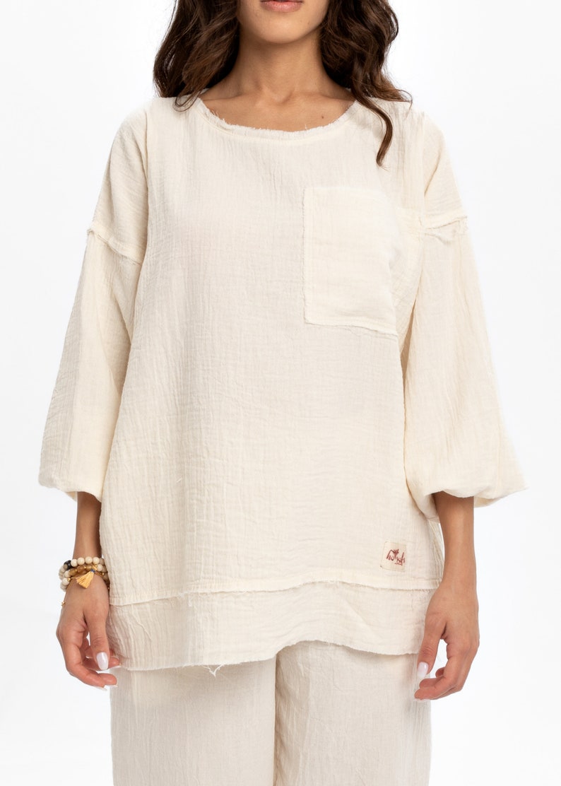 May include: A white, loose-fitting, 3/4 sleeve top with a small pocket on the left side. The top has a raw edge hem and a small label with the word "Nude" on the bottom right side.