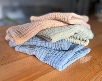 May include: A stack of four soft, textured fabric squares in shades of light blue, green, and beige. The fabric is folded and arranged on a wooden surface.