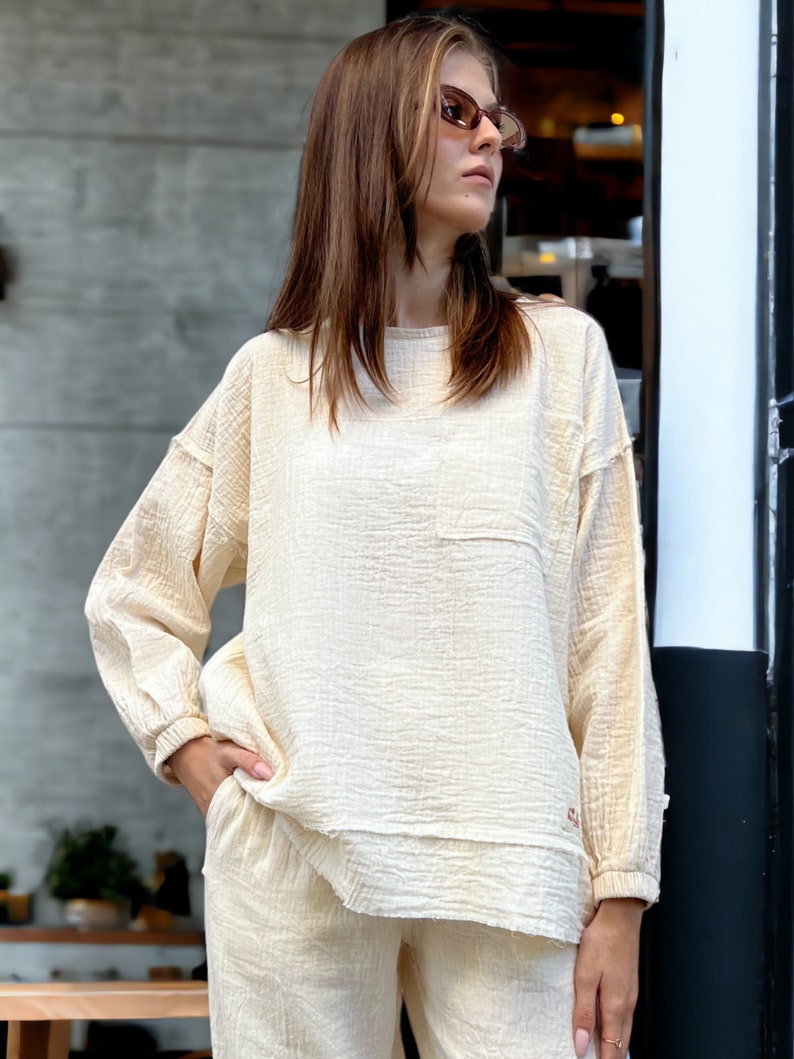 May include: A cream-coloured, long-sleeved, loose-fitting top with a small pocket on the left side. The top is made of a soft, textured fabric and has a slightly frayed hem. The model is wearing the top with matching trousers.