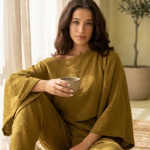 May include: A two-piece lounge set in olive green, featuring a loose-fitting top with wide, flared sleeves and a wide neckline, paired with matching relaxed-fit trousers. The fabric has a visible crinkled texture throughout the entire garment.