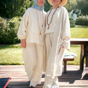 May include: Two people wearing white linen clothing. One person is wearing a blue headscarf and a long necklace. The other person is wearing a wide-brimmed straw hat and a shorter necklace. They are standing on a wooden deck.