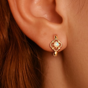 Può includere: Un orecchino a cerchio dorato con un design floreale a quattro petali e una piccola pietra rotonda e opalescente al centro. L'orecchino è indossato su un orecchio, evidenziando gli intricati dettagli del gioiello e la sottile lucentezza della pietra.