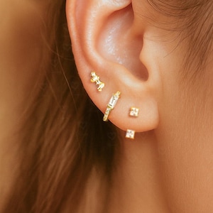 Könnte beinhalten: Nahaufnahme eines Ohrs, das mit mehreren goldenen Ohrringen geschmückt ist. Die Ohrringe umfassen einen kleinen Stecker, einen Reifen mit einem rechteckigen Edelstein und ein Cluster-Design. Der Schmuck ist zart und verleiht einen Hauch von Eleganz.