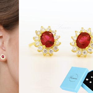 Może przedstawiać: Para złotych kolczyków z czerwonym owalnym kamieniem szlachetnym otoczonym mniejszymi białymi kamieniami szlachetnymi. Kolczyki znajdują się w niebieskim pudełku z napisem "Leswin" na nim.