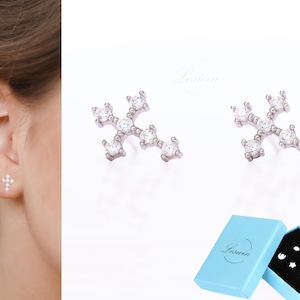 May include: A pair of silver snowflake earrings with clear cubic zirconia stones. The earrings are shown in a close-up view and are packaged in a blue box with the brand name "Leswin" printed on it.